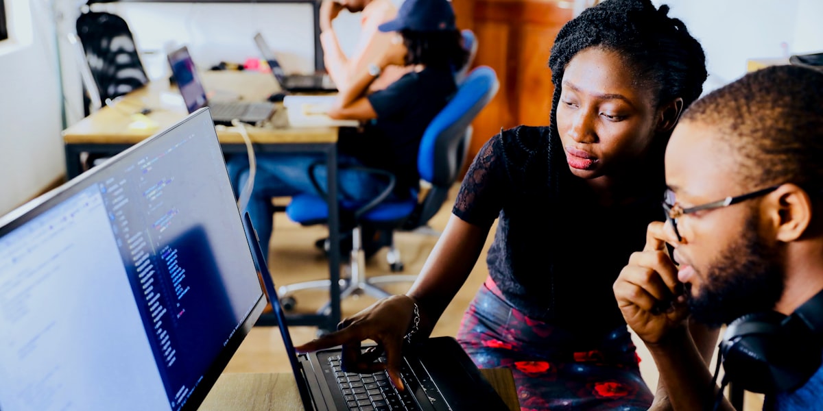 Two colleagues reviewing code together on a monitor, representing AI adoption raising the skill floor and reskilling needs