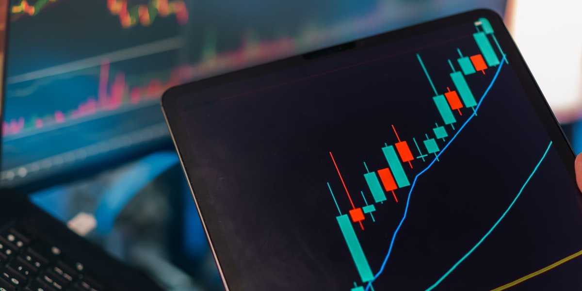 Hand holding a tablet displaying a rising candlestick market chart, with additional trading screens visible in the background, representing the week's synthesis of market signals